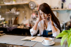 Stressed business owner at counter, with financial books, seeking advice on phone.